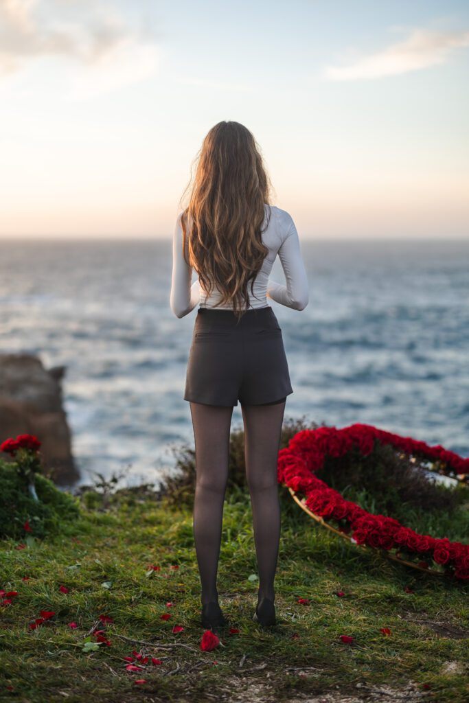 A woman with long hair stands on grass facing the ocean at sunset, wearing a white top, black shorts, and black tights. Red roses and a heart-shaped arrangement lie beside her—a perfect scene for a Big Sur engagement photographer.
