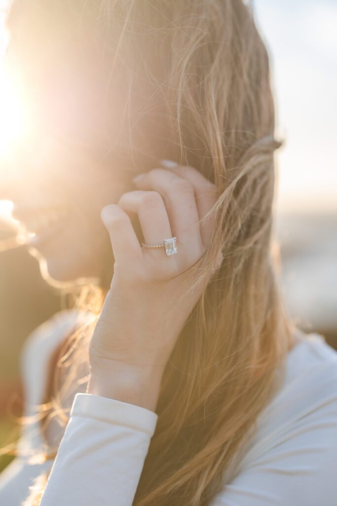 A woman with long blonde hair smiles in soft sunlight, holding her hand to her face and displaying a sparkling rectangular-cut engagement ring—perfectly captured by a Carmel proposal photographer.