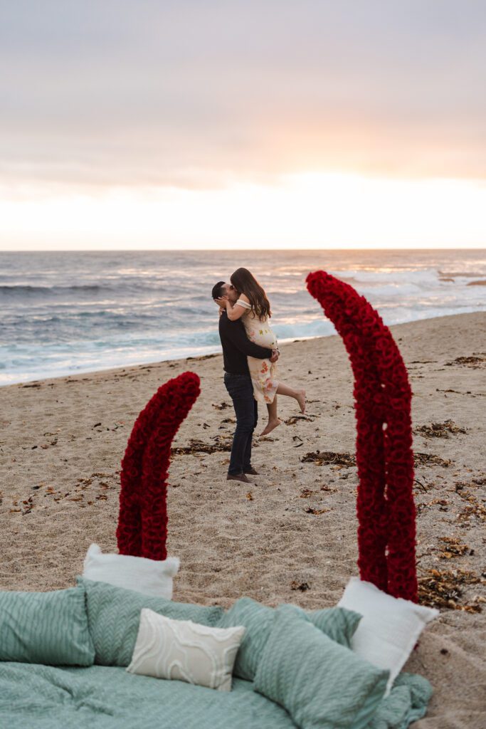 A man lifts a woman in an embrace on a sandy beach at sunset. In the foreground, a bed with green bedding and red rose-decorated tentacles sits by the shoreline, perfect for a romantic Carmel by the Sea proposal captured by a skilled carmel photographer.