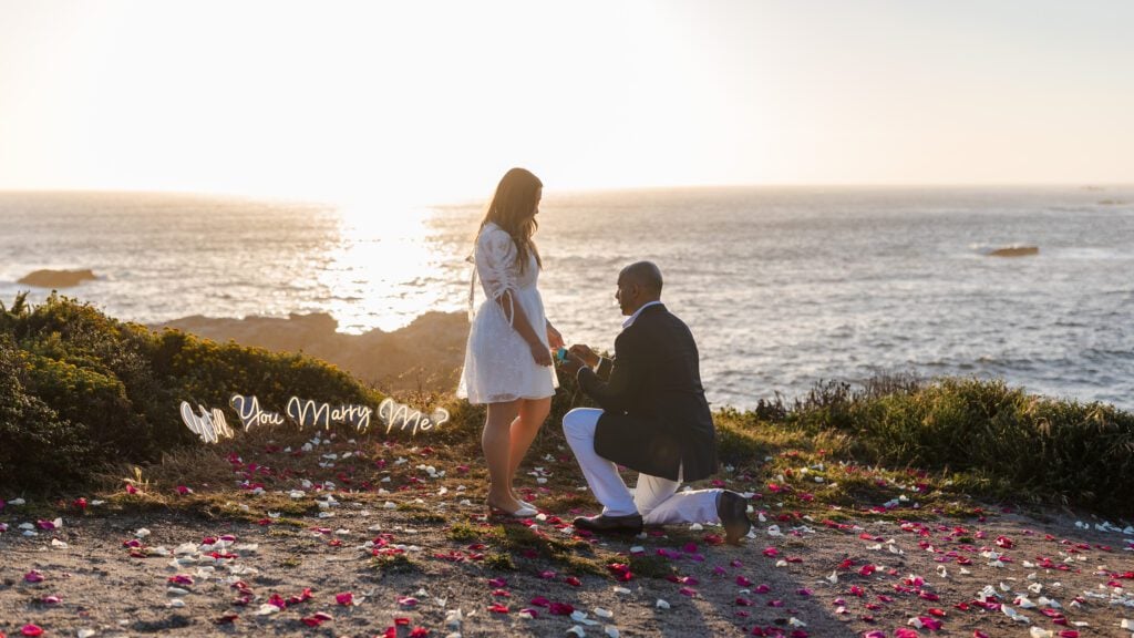 A man kneels and proposes to a woman at sunset by the ocean, with rose petals scattered on the ground and Will you marry me? written in white script nearby—a magical Big Sur proposal captured by a talented Carmel photographer.