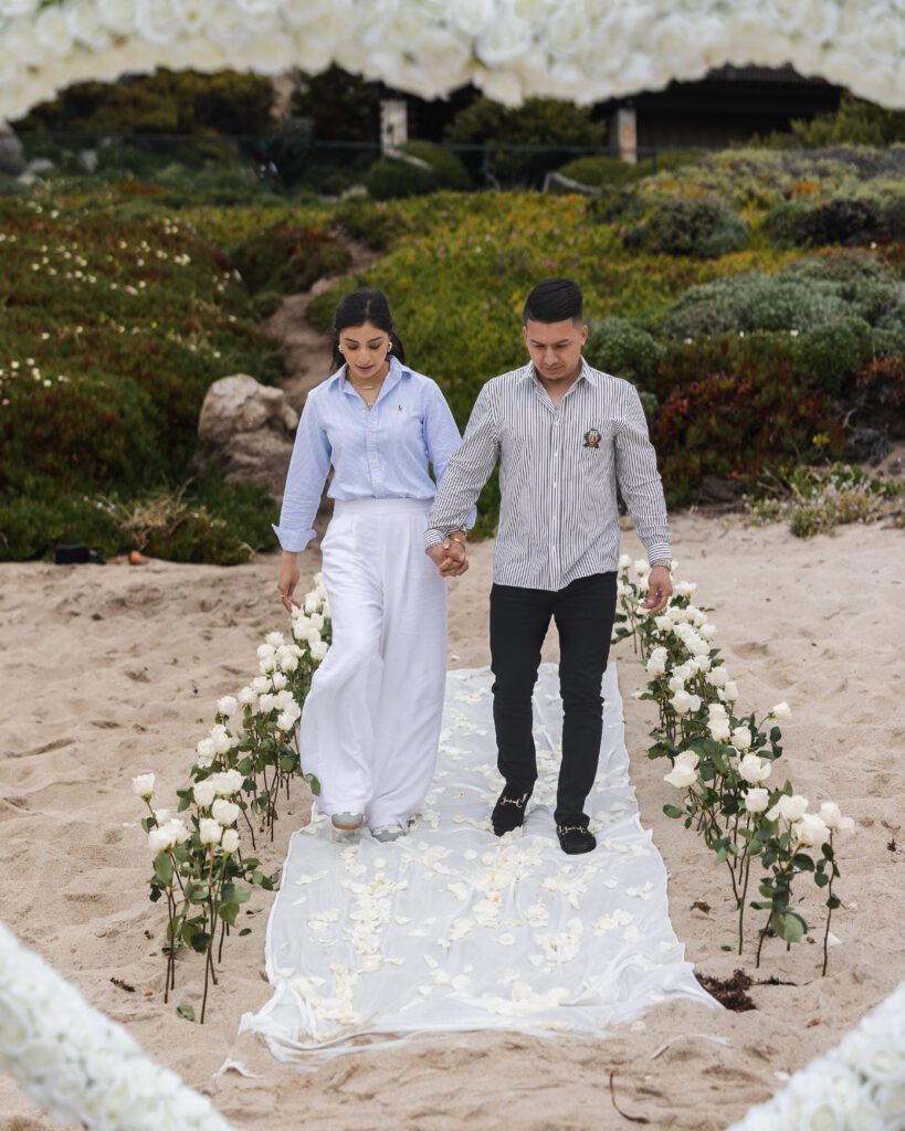 A couple walks hand-in-hand along a white rose-lined aisle on a sandy beach, stepping on a fabric runner covered with rose petals—an unforgettable moment captured by a Carmel photographer, surrounded by lush greenery.