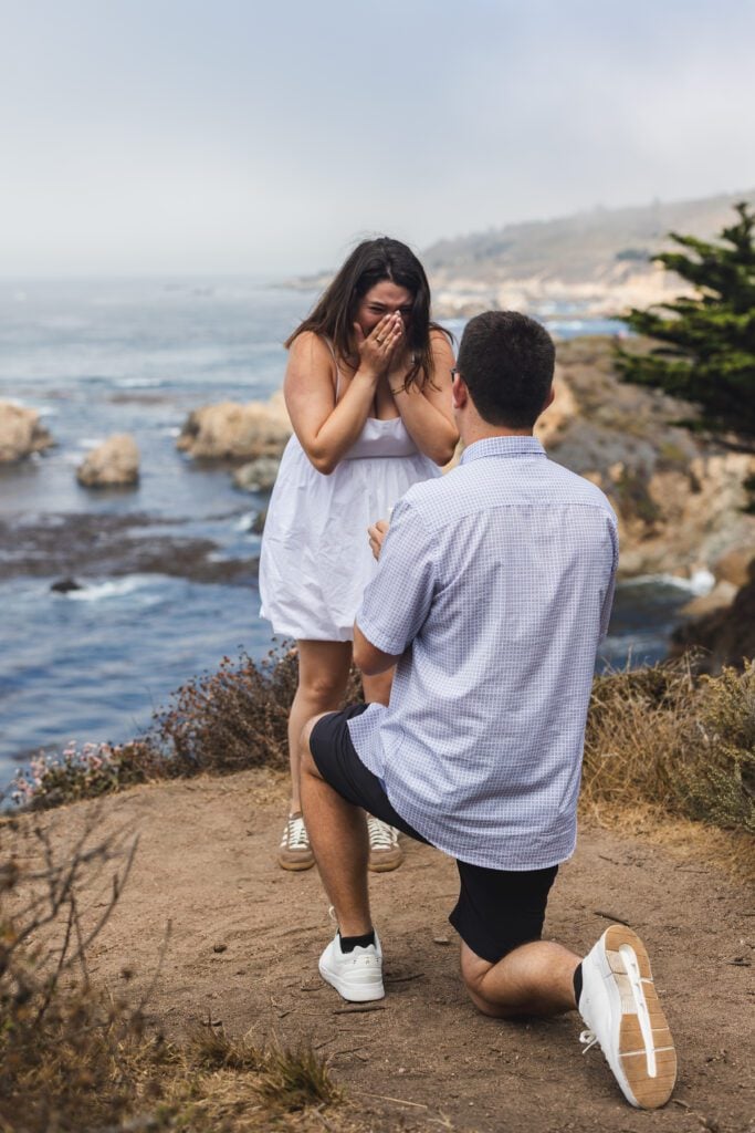 A man kneels and proposes to a surprised woman by the seaside, with rocky cliffs and ocean waves in the background—a magical Carmel by the Sea proposal captured as she covers her face with her hands in excitement.