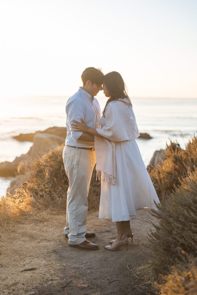 A couple stands close together on a coastal cliff at sunset, gently touching foreheads. Captured by a Big Sur engagement photographer, they wear light, summery clothes as ocean waves and rocky views frame their romantic moment.