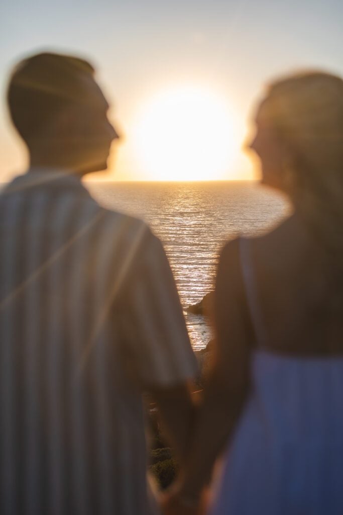 A couple holding hands is seen from behind, standing together and looking at the sunset over the ocean. Sunlight reflects on the water, creating a warm, romantic atmosphere—perfect for a Big Sur engagement photographer to capture.
