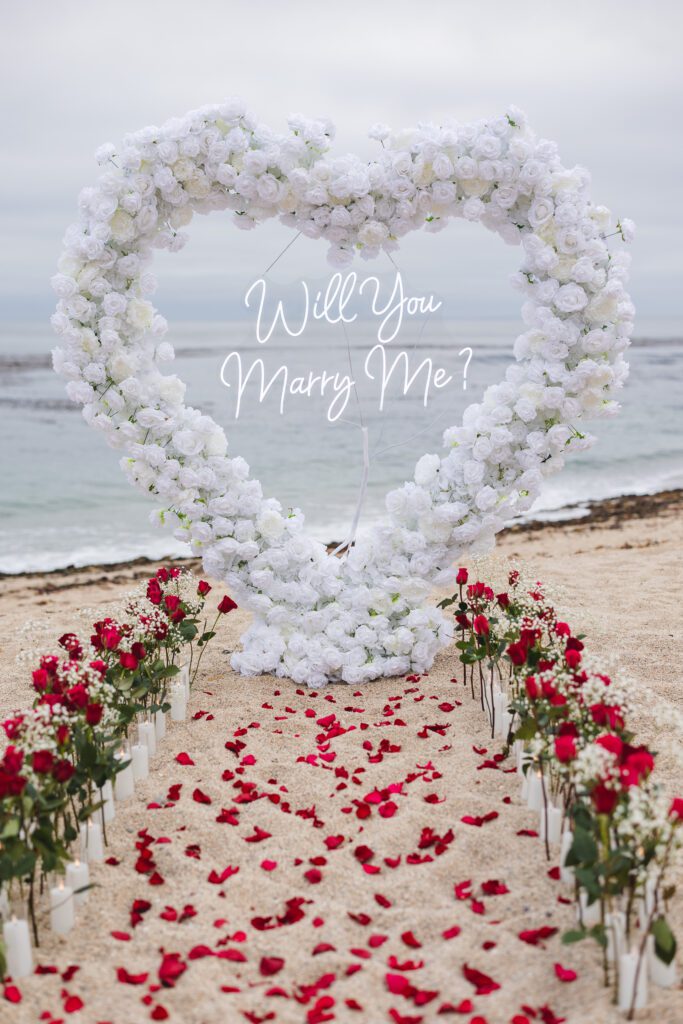 A heart-shaped arch made of white flowers stands on a sandy beach, framed by red roses and rose petals. “Will You Marry Me?” appears in script at the center—an idyllic Carmel by the Sea proposal with the ocean as your backdrop.