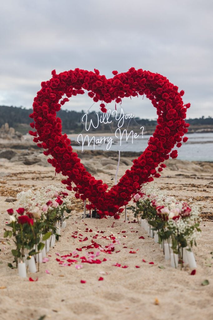 A large heart-shaped arrangement of red roses with a “Will you marry me?” sign stands on a sandy beach, surrounded by flower vases and rose petals—a dreamy Carmel by the Sea proposal captured perfectly for your special moment.