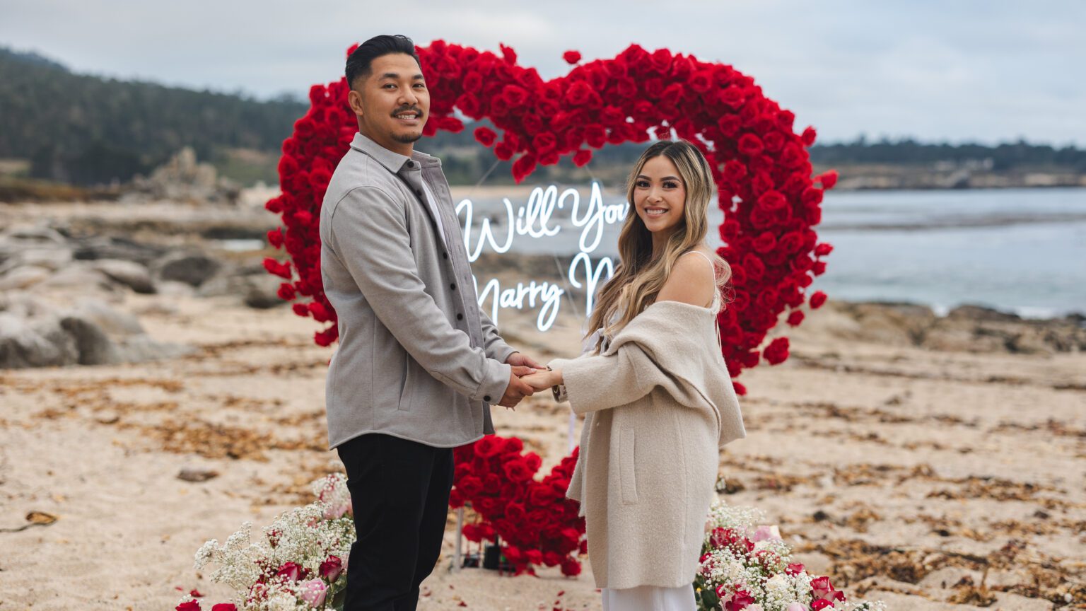A couple holding hands and smiling on a beach in Carmel by the Sea, standing before a heart-shaped arrangement of red roses and a neon Will You Marry Me? sign. Flowers are scattered around them, perfect for capturing by a Carmel proposal photographer.