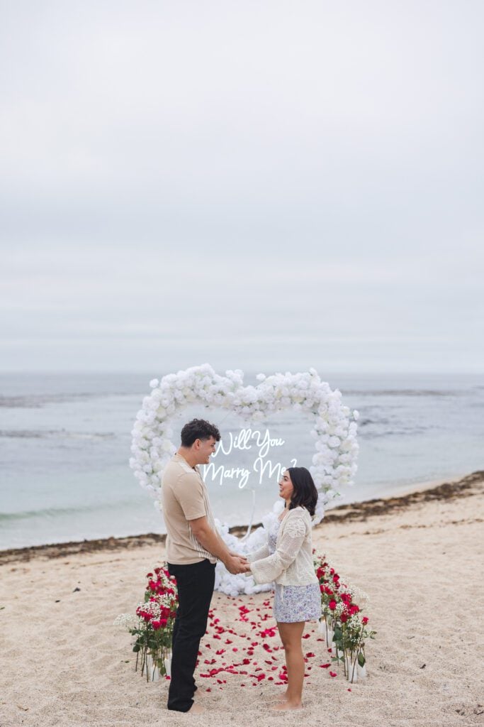 A couple stands on a beach holding hands in front of a heart-shaped flower arch and a “Will You Marry Me?” sign, surrounded by roses and red petals on the sand—captured perfectly by a Big Sur proposal photographer with the ocean in the background.