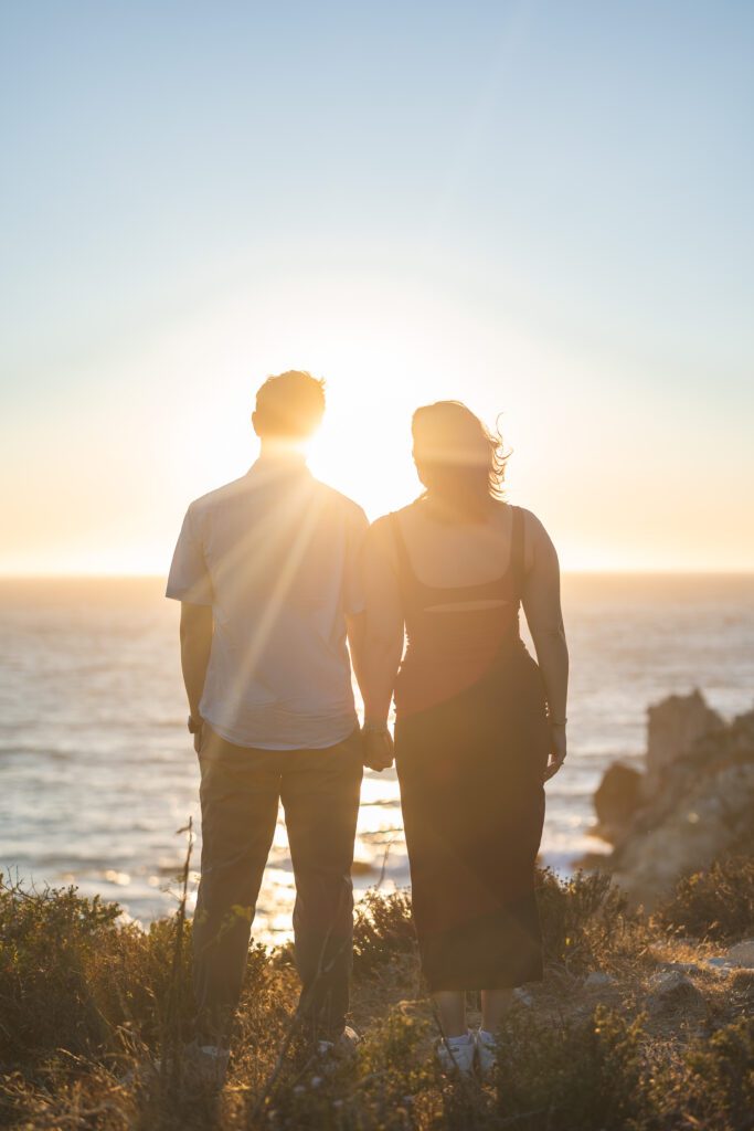 A couple stands hand in hand, facing the ocean at sunset. The sunlight creates a warm glow as they look out over the water—an unforgettable moment captured by a Carmel photographer, with lush plants visible in the foreground.