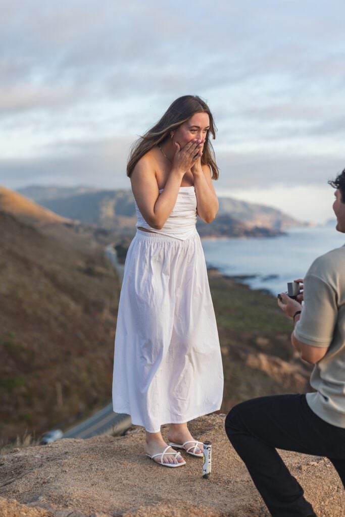 A woman in a white dress stands on a hilltop, covering her mouth in surprise, while a man kneels with an open ring box—a breathtaking Big Sur proposal with mountains and water in the background.