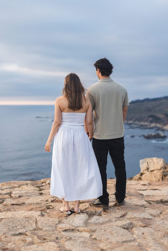 A woman in a white dress and a man in a light shirt and dark pants stand side by side on a rocky cliff, looking out at the ocean under a cloudy sky—a classic scene for any Big Sur engagement photographer.