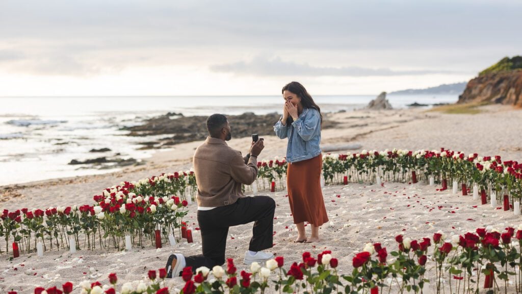 A man kneels and proposes to a surprised woman on a beach, surrounded by red and white roses arranged in a heart. The ocean and shoreline glisten behind them, perfectly captured by a Big Sur proposal photographer.