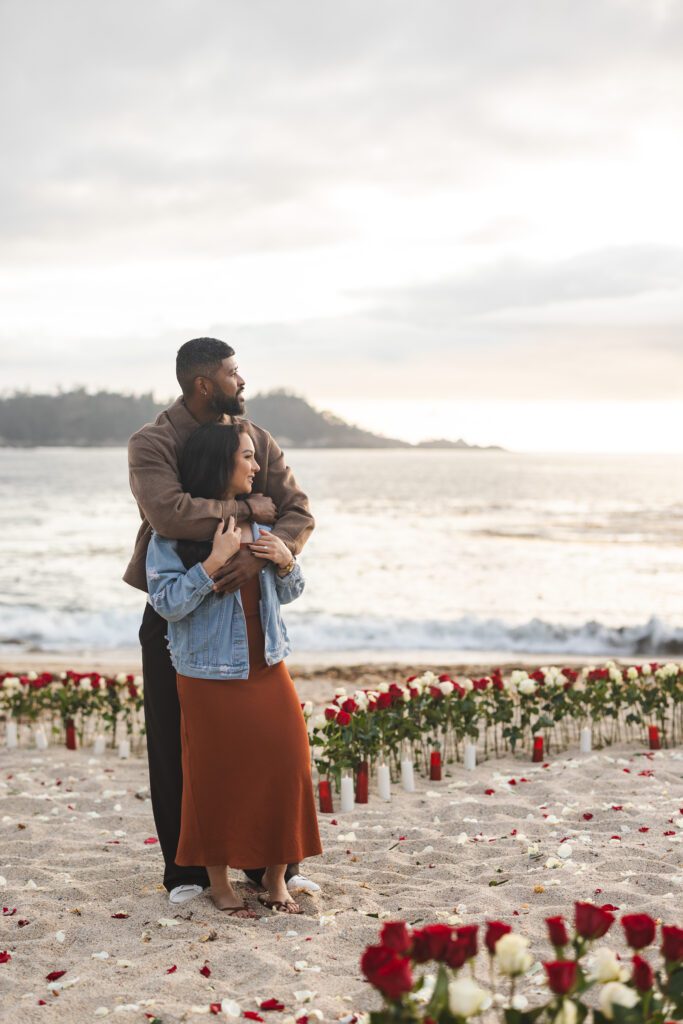 A couple stands on a beach at sunset, embracing each other. Surrounded by rows of red and white roses, they celebrate their love as a Big Sur proposal photographer captures the moment against the ocean and distant hills.