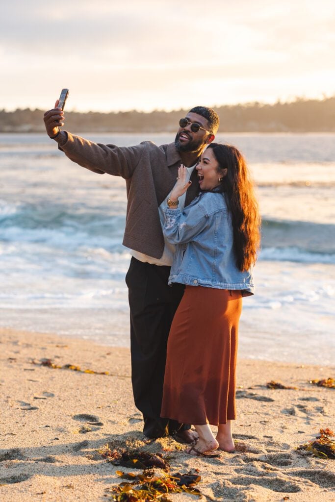 A smiling couple stands barefoot on a sandy beach near the ocean, taking a selfie together at sunset. The man wears sunglasses and a sweater, while the woman wears a denim jacket and a rust-colored skirt.