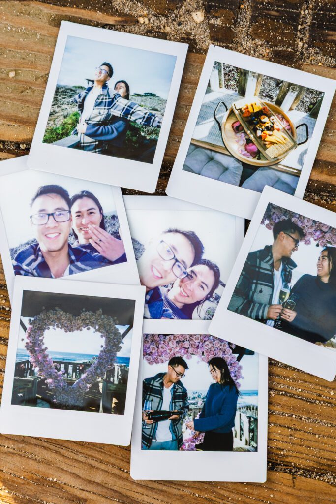 A collection of seven Polaroid photos arranged on a wooden surface, capturing a couple posing together, sharing drinks, enjoying food, and standing near heart-shaped floral decorations by a Big Sur proposal photographer.
