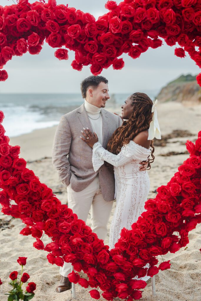 A couple stands on a beach, smiling at each other inside a large heart-shaped frame made of red roses. The woman wears a white lace dress; the man wears a light suit. Waves and a rocky shoreline are visible in the background.