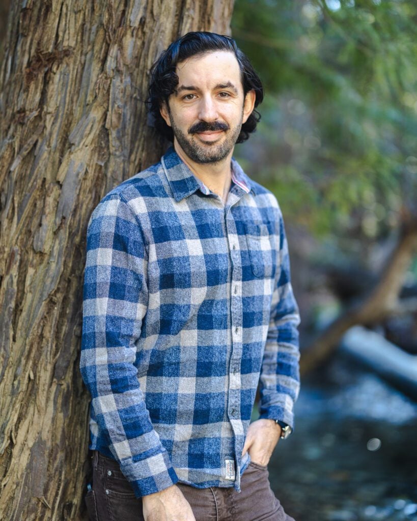 A man with dark hair and a mustache, wearing a blue and gray plaid shirt, stands outdoors leaning against a tree, evoking the relaxed charm of a Big Sur proposal with a blurred natural background.