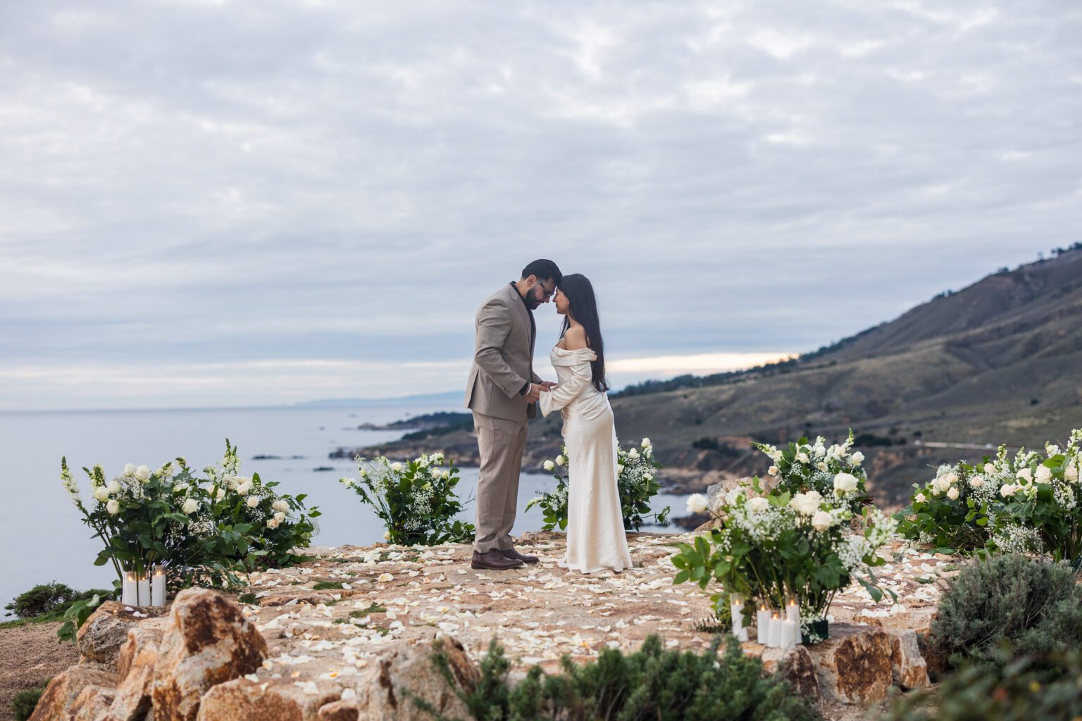 A couple stands close together on a rocky cliff decorated with white flowers and candles, overlooking the ocean and rugged coastline under a cloudy sky—a perfect scene for a Big Sur proposal photographer to capture.