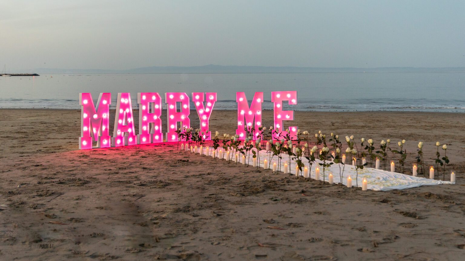 Large illuminated Marry Me letters on a sandy beach, with a white aisle lined with candles and white roses leading to the sign; calm sea and sky in the background—perfect for capturing by a Big Sur proposal photographer.