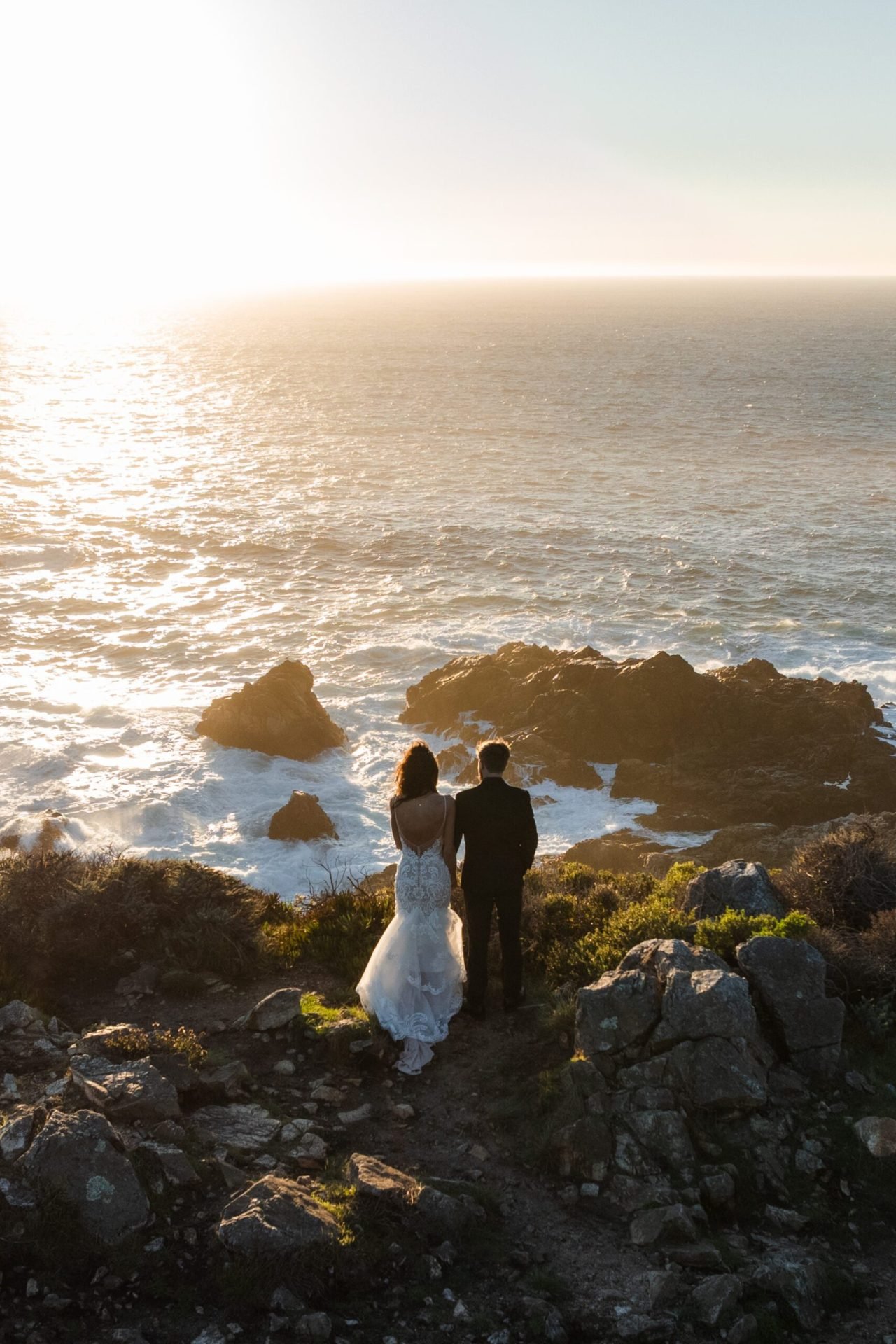 A bride and groom stand on a rocky cliff, holding hands and looking out at the ocean as the sun sets—a moment perfectly captured by a Big Sur proposal photographer, with a warm glow over the water and landscape.